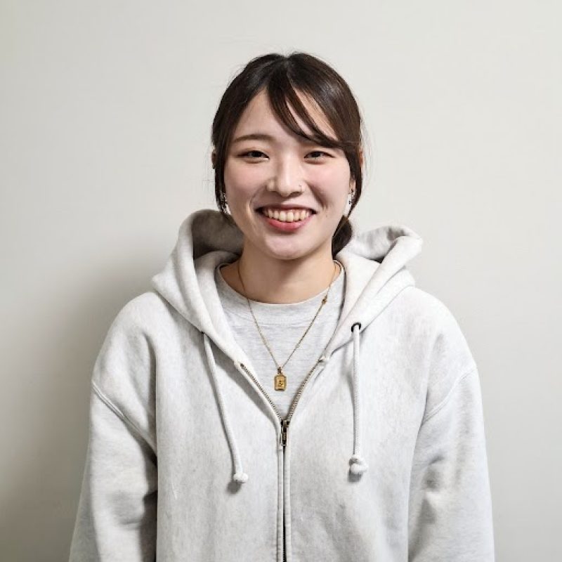 Smiling young woman in a light gray zip-front hoodie, standing against a pale wall.